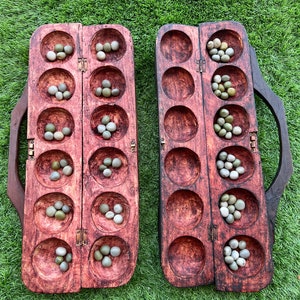 May include: A wooden game board with two rows of six holes, each containing small, round, green and white stones. The board is made of dark wood and has a reddish-brown stain. The board is open and ready for play.