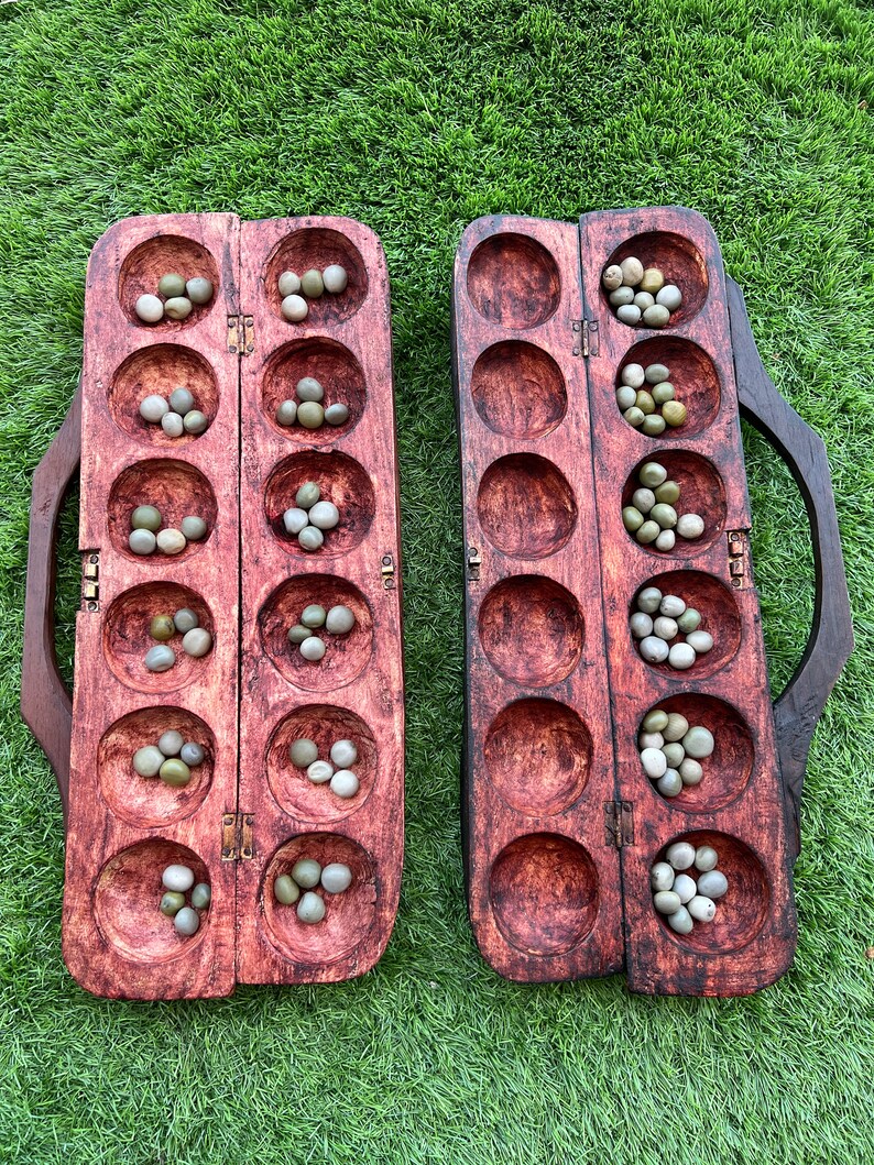 May include: A wooden game board with two rows of six holes, each containing small, round, green and white stones. The board is made of dark wood and has a reddish-brown stain. The board is open and ready for play.