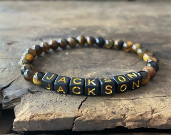 Puede incluir: Una pulsera de cuentas con piedras ojo de tigre redondas y lisas en tonos marrón y dorado, intercaladas con cuentas cuadradas negras. Las cuentas negras tienen impresas letras doradas que forman el nombre JACKSON en secuencia.