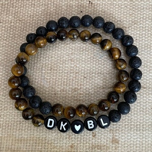May include: Two bracelets, one with black lava beads and the other with tiger's eye beads. The bracelets are connected with letter beads that spell "DK" and "BL" with a heart bead in between.
