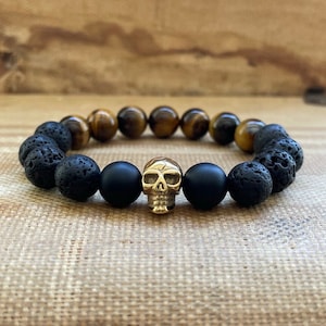 May include: A bracelet made with black lava rock and matte black beads, featuring a gold skull charm and tiger's eye beads.