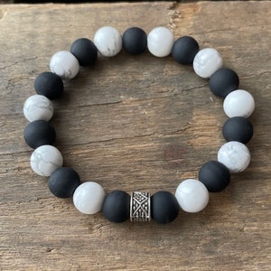 May include: A black and white beaded bracelet with a silver charm. The bracelet is made of round beads with a matte finish.