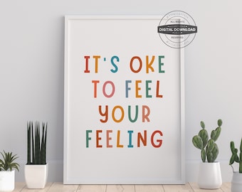 May include: A white framed print with the text "It's okay to feel your feeling" in colorful letters. The print is on a white wall with a wooden floor and three potted plants.
