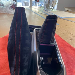 May include: Black and red car interior gear shift and parking brake boot covers. The covers are made of a black fabric with red stitching.