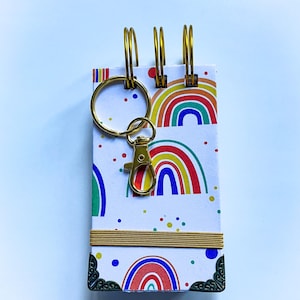 May include: A small notebook with a rainbow and polka dot pattern. The notebook has a gold ring binding and a gold key chain attached.