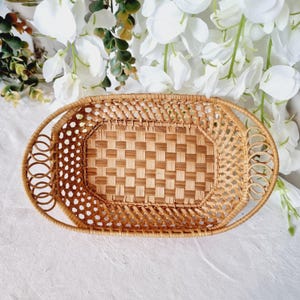 Puede incluir: Cesta ovalada de mimbre tejido, con base de patrón a cuadros y asas decorativas. La cesta marrón claro tiene un aspecto natural y rústico. Las asas presentan un diseño en bucle. La cesta está vacía y sobre una superficie blanca.