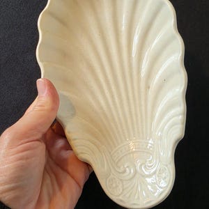 Könnte beinhalten: Cremefarbene, muschelförmige Schale mit gewelltem Rand. Die Keramikschale hat ein detailliertes, geriffeltes Design und dekorative Schnörkel am Boden. Die Schale wird vor einem schwarzen Hintergrund gehalten.