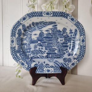 May include: A blue and white porcelain serving platter with a landscape design. The octagonal platter features a detailed scene of buildings, trees, and mountains. The rim has a decorative border. The platter is displayed on a wooden stand.