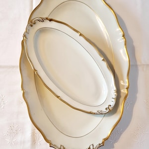 May include: Two white oval serving platters with gold trim and decorative edges. The platters are nested, with a smaller one on top of a larger one. The gold trim has a delicate, ornate design. The background is a white tablecloth.