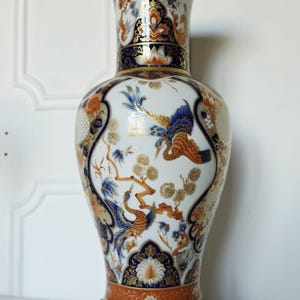 Può includere: Un vaso in ceramica decorato con motivi di uccelli e fiori nei toni del blu, arancione e oro. Il vaso è posto su un supporto in legno scuro. L'altezza del vaso è di circa 40 cm.
