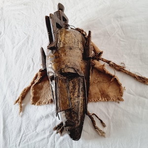 May include: A handcrafted, elongated mask with a dark brown finish. The mask features intricate metalwork and is adorned with wooden elements and fabric accents. The design includes a figure at the top and braided rope details, showcasing a tribal aesthetic.