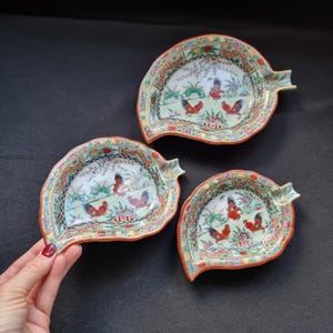 Puede incluir: Tres platos de porcelana con forma de hoja y motivo de gallo. Los platos tienen un borde rojo y una escena interior detallada con gallos, flores y árboles. Los platos son de diferentes tamaños.