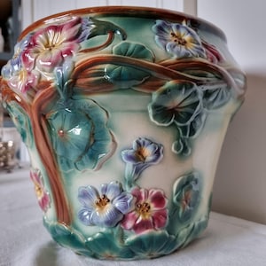 May include: A ceramic flower pot with a detailed relief design. The pot features raised floral and leaf motifs in shades of blue, purple, pink, and green, with brown accents. The pot has a glossy finish.