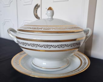 May include: White porcelain soup tureen with a lid, handles, and a matching saucer. The tureen and saucer are decorated with gold trim and a black and gold floral pattern. The lid has a gold finial.