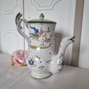 Op de afbeelding: Witte keramische kan met deksel, versierd met een vogel- en bloemmotief in blauw, oranje en groen. De kan heeft een unieke tuit en handvat, met een groen accent op het deksel. Het ontwerp doet denken aan vintage aardewerk.