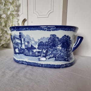 May include: A blue and white ceramic planter with a landscape design. The planter has a rectangular shape with rounded edges and two handles. The landscape scene depicts buildings, trees, and figures. The rim and handles are a darker blue.