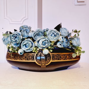 Può includere: Una fioriera in ceramica blu scuro con filigrana dorata e un emblema centrale, riempita di rose artificiali blu chiaro e verde. Il contenitore di forma ovale è un elemento decorativo.