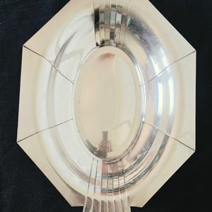 Op de afbeelding: Een gepolijst zilverkleurig metalen dienblad met een achthoekige buitenkant en een centrale ovale komvorm. Het dienblad is voorzien van decoratieve verticale gegroefde lijnen aan de boven- en onderrand, wat zorgt voor een gestructureerd ontwerp.