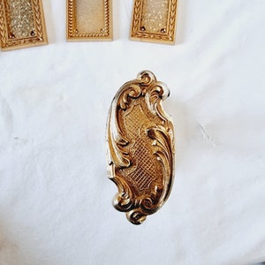 Peut inclure: Un fermoir en métal doré orné d'un motif de volutes détaillé. Le fermoir a une surface texturée et à motifs et est entouré d'éléments décoratifs. Trois petits cadres dorés sont visibles en arrière-plan.