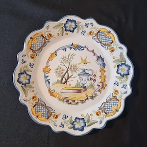 Peut inclure: Une assiette en céramique blanche à bord festonné, ornée d'un motif peint à la main. Le motif comprend des motifs floraux bleus et jaunes, une scène centrale avec un vase, un arbre et un oiseau, et une bordure bleue. L'assiette mesure environ 30 cm de diamètre.
