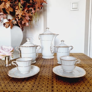 May include: A white porcelain tea set with gold trim, including a teapot, creamer, sugar bowl, and two teacups with saucers. The set is arranged on a patterned brown tablecloth, with decorative elements in the background.
