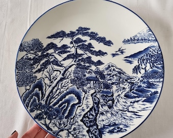 May include: White ceramic plate with a blue landscape design. The plate features a detailed illustration of a mountain scene with trees, a river, and a small building. The rim of the plate has a thin blue border.