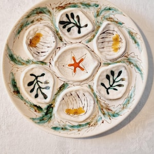 May include: A white ceramic oyster plate with six individual wells and a central well. The plate is decorated with hand-painted designs, including a starfish, leaves, and floral accents in orange, black, and green.