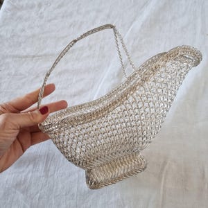 May include: A silver-toned wire basket with a handle. The basket has a woven design and a curved shape, with a flared base. The handle is made of twisted wire and chain. The basket is held by a hand.