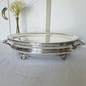 Puede incluir: Una fuente plateada con tapa y asas. La fuente ovalada se apoya sobre patas decorativas y tiene una superficie reflectante. Un pequeño jarrón con flores blancas está en el fondo.