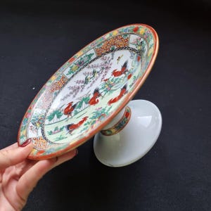 Peut inclure: Un plat de service ovale et sur pied avec un design coloré et détaillé. Le plat présente un bord rouge et doré, avec une image centrale d'oiseaux rouges, de fleurs et de verdure. La base est blanche avec une bande décorative assortie.