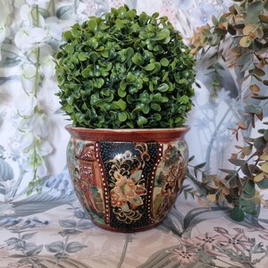 Peut inclure: Un pot de fleurs décoratif en céramique avec un motif coloré et détaillé représentant des personnages et des paysages. Le pot est rempli d'une plante verte et feuillue. Le bord du pot est rouge foncé.