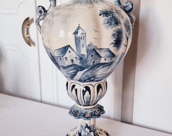 May include: A tall, ornate ceramic vase with a blue and white color scheme. The vase features a landscape scene with buildings and trees. It has dragon-shaped handles and a decorative base, perfect for home decor.