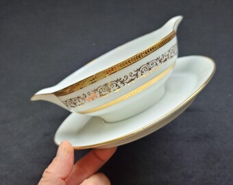 May include: White porcelain gravy boat with a matching saucer. The gravy boat and saucer are trimmed with gold bands and decorative gold filigree patterns. The gravy boat has a spout and a handle. The set is on a dark background.