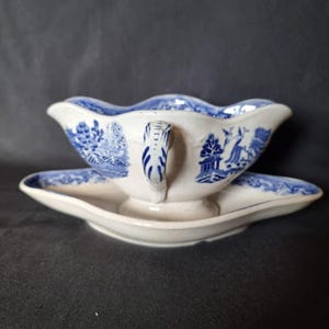 Può includere: Salsiera in ceramica bianca con un disegno a paesaggio blu e bianco. La barca ha un manico e poggia su un piattino abbinato. Il bordo della barca e del piattino è smerlato.