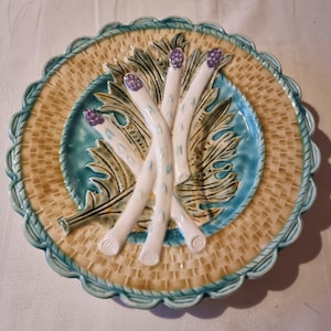 Puede incluir: Un plato de cerámica decorativo con un diseño central de espárragos blancos con puntas moradas, sobre un patrón de hojas verde y turquesa. El plato tiene una textura tejida en beige y un borde festoneado turquesa.