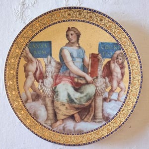 Peut inclure: Assiette décorative ronde avec un bord doré et une image centrale d'une femme en robe bleue tenant un livre. L'assiette présente des chérubins et du texte en latin. Le fond est doré avec une bordure bleue.