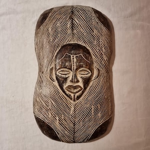 Op de afbeelding: Een gesneden houten masker met een centraal gezichtsontwerp. Het masker heeft een donkerbruin gezicht met gebeeldhouwde gelaatstrekken, omgeven door een lichtere bruine, getextureerde achtergrond. Het masker is ongeveer 46 cm hoog en 25 cm breed.