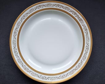 May include: A white porcelain plate with a gold rim and decorative gold filigree border. The plate has a slightly recessed center and is set against a dark background. The gold detailing adds a touch of elegance to the tableware.