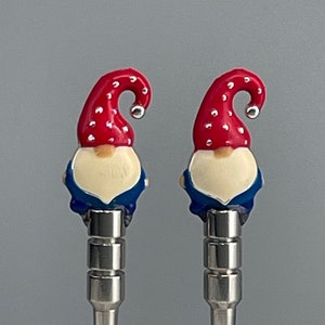 May include: Two decorative bottle stoppers featuring gnome designs. Each gnome has a red hat with white polka dots, a cream-colored face, a blue jacket, and a silver metal stem. The stoppers are set against a gray background.