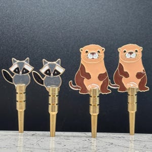 May include: Four decorative metal plugs with animal designs. Two plugs feature raccoons with black and white details, and two have otters in brown and tan. The plugs have a gold-colored base.