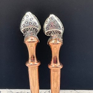 May include: Two decorative bottle stoppers with mushroom-shaped tops. The mushroom caps are silver with small, evenly spaced circles. The stems are copper-colored, with a cylindrical shape and a flared base.