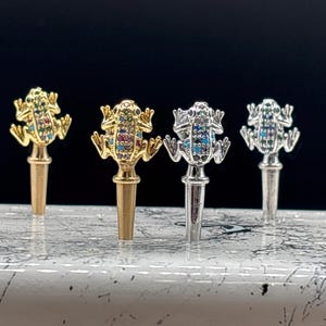 May include: Four bottle stoppers with frog designs. The frogs are made of metal and have colorful enamel details. Two of the stoppers are gold, one is silver, and one is a light gold color.