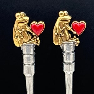May include: Two gold frog figurines holding red hearts. The frogs are sitting on silver metal rods.