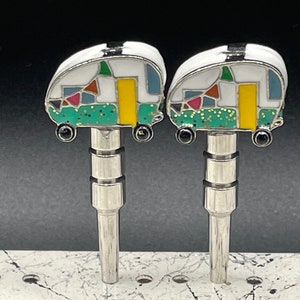 May include: Two silver metal bottle stoppers with a white camper design. The camper is decorated with green, yellow, red, blue, and black enamel paint. The camper has a sparkly green roof.
