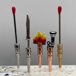 May include: Five miniature metal torch-like objects with different designs. The objects are silver, gold, and copper colored. One object has a red and yellow flame design. The objects are on a white surface.