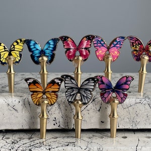 May include: Eight decorative butterfly bottle stoppers with gold metal bases. The butterflies are painted in various colors, including yellow, blue, pink, orange, white, and purple.