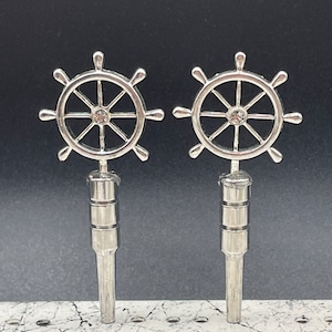 May include: Two silver-toned ship wheel earrings with a clear rhinestone in the center of each wheel. The earrings are on a white surface with black markings.