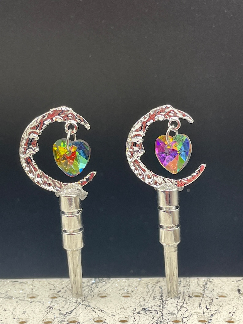 May include: Two silver crescent moon charms with iridescent heart-shaped crystals hanging from them. The charms are attached to silver posts.