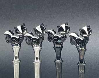 May include: Four silver and black metal bottle stoppers shaped like skunks. The stoppers have a shiny finish and are in a row on a white marble surface.