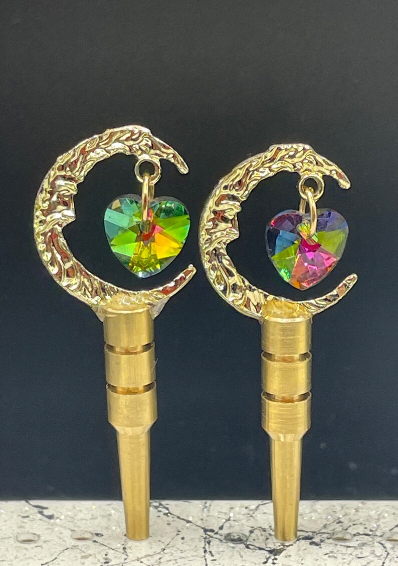 May include: A pair of gold-tone earrings with a crescent moon design and a dangling heart-shaped crystal. The crystals are iridescent and have a rainbow of colors.
